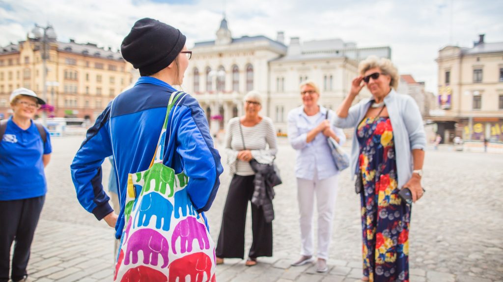 Sinitakkinen opas seisoo Keskustorilla selin kameraan elefanttikuvioinen kassi olallaan.