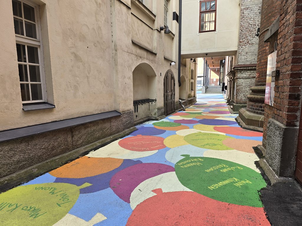Finlaysonin alueen katumaalaus kesältä 2024. Kapealla käytävällä vaaleankeltaisten ja punatiilisten seinien välissä asvalttiin maalattuja monivärisiä ilmapalloja, joihin on kirjoitettu taiteilijoiden nimiä.