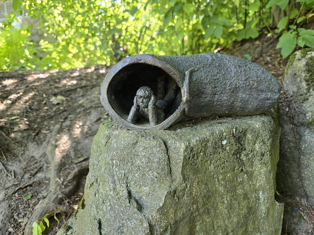 Kiven päällä pieni pronssipatsas, joka esittää kengästä kurkistelevaa ihmistä.